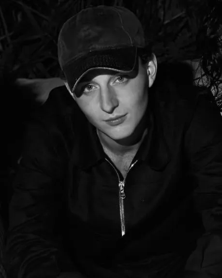 Black and white portrait of a man wearing black hoodie, cap, smiling into the camera, artist portrait of Dutch American sculptor levi de jong, impulse magazine interview.