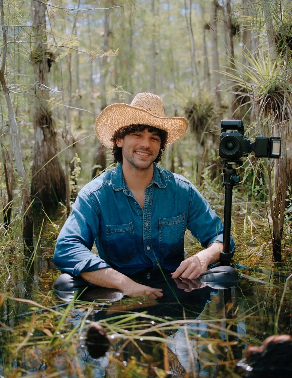 A man in denim shirt standing in swamp water with camera and straw hat smiling, portrait of photographer josh aronson at work, impulse magazine interview.