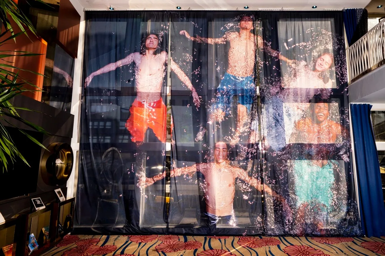 Installation view of photograph of florida boy bathing under sunlight with eyes closed printed on translucent fabric hanging on window of catalina hotel lobby, miami beach, josh aronson commission at no vacancy 2024, impulse magazine interview.