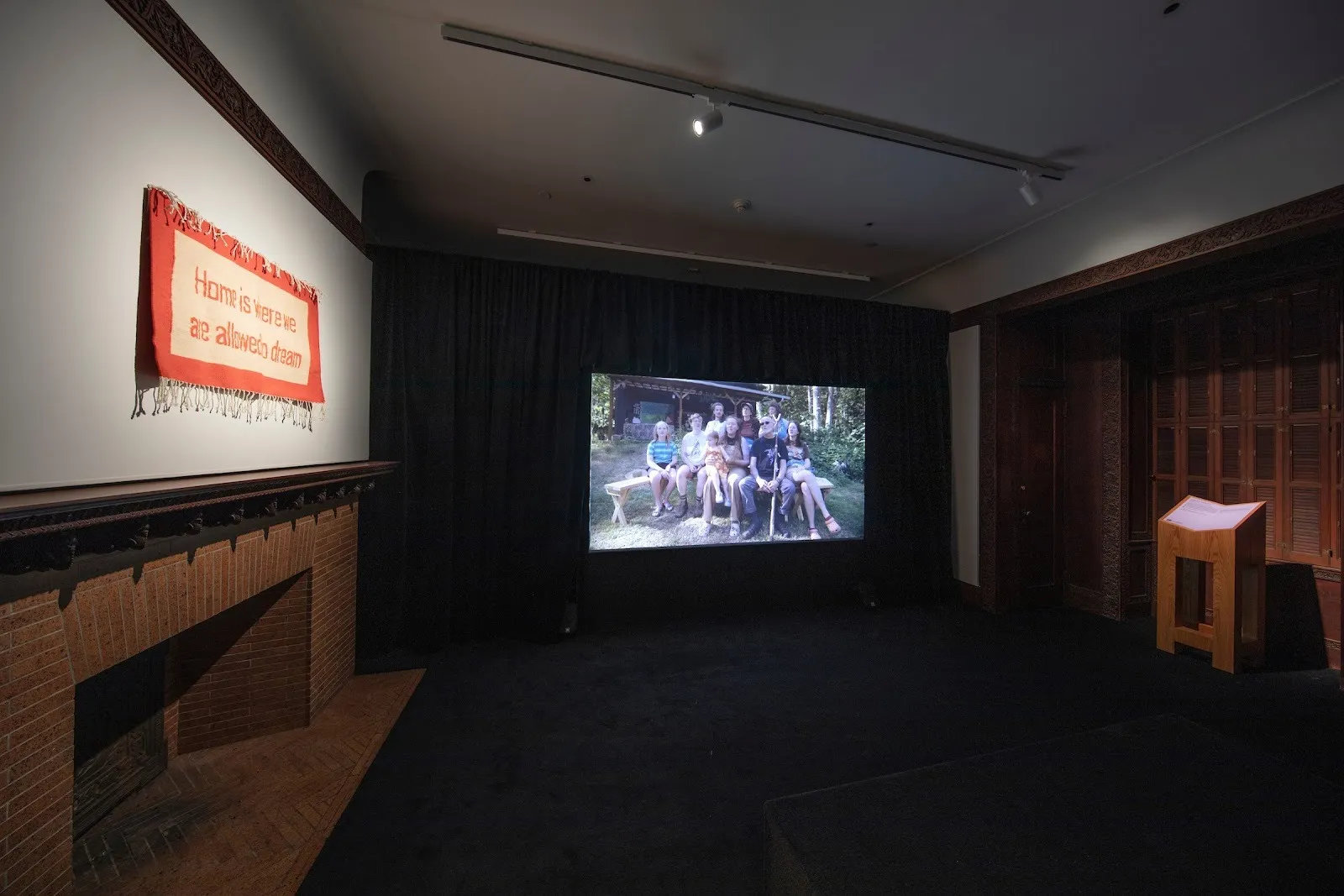 Museum room installed with projector, textile saying home is where we are allowed to dream, documentary of trans Community center in Arkansas, Smithsonian Museum design triennial making home sterling corum review.