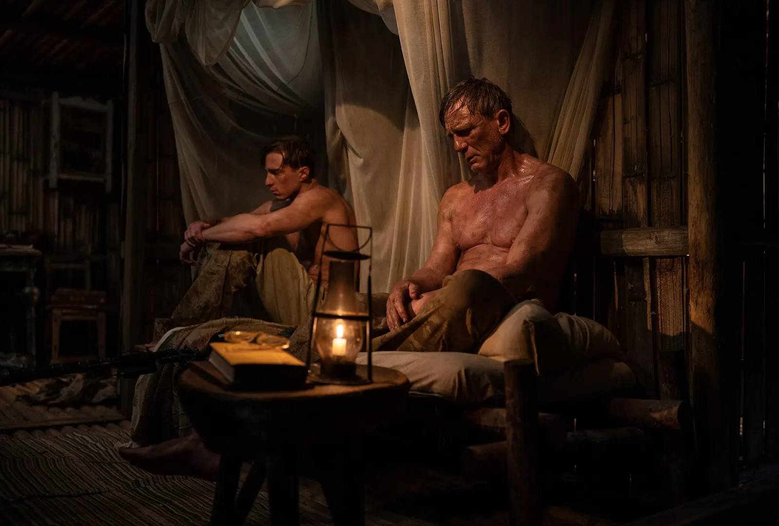 Two shirtless men sit in a dimly lamp-lit shack draped with brown sheets. They sit against the wall, hunched and slouched, looking dejected. The man on the right rests his forearms on his knees drawn. The man on the right rests his hands in his lap.