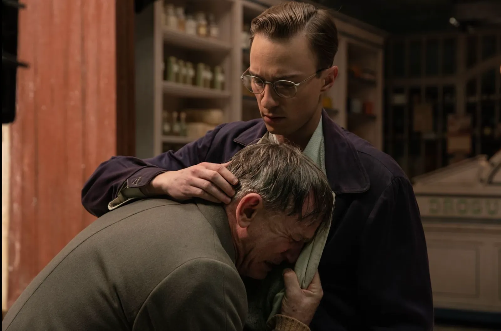 Two men embrace each other as the man on the right is hunched over, gripping and crying into the jacket and shirt of the man on the right. The man on the right, wearing glasses, comforts the other by placing his hand on the crying man's shoulders.