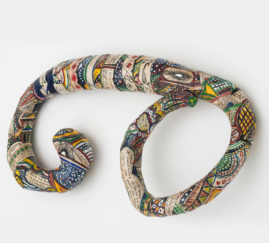 A thin, snake-like sculpture curls in on itself on either end, creating a deeply curved form. On the surface of the sculpture, a collage of text and colorful hand-painted designs and symbols are pasted on and covers the surface.