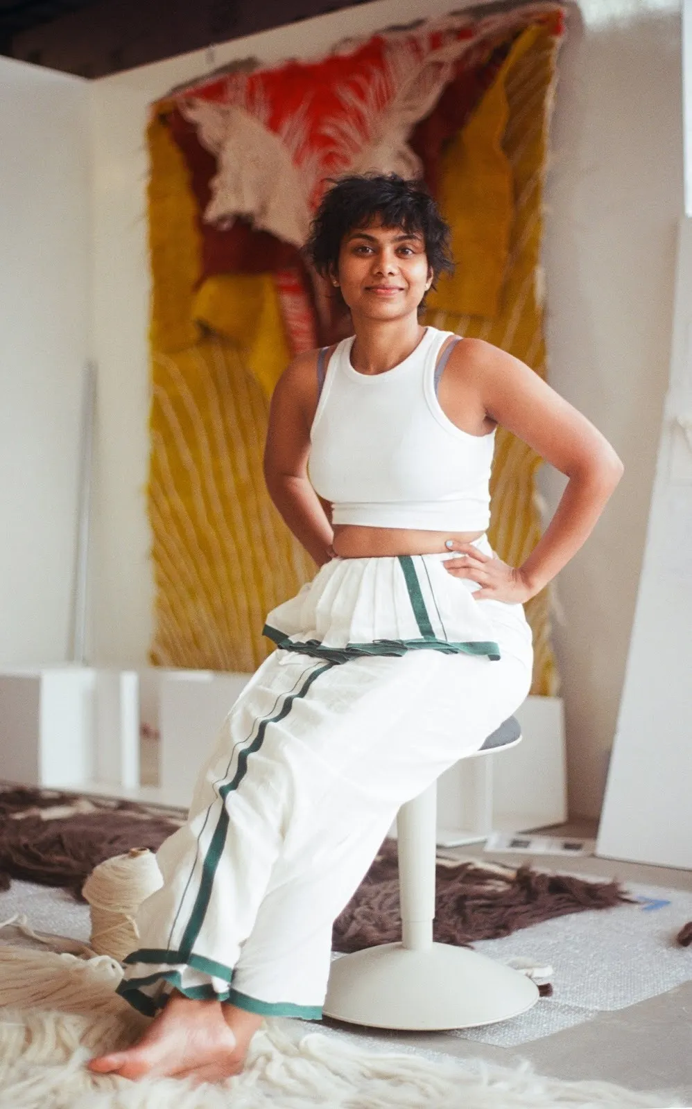 Artist Sagarika Sundaram sitting on stool in white clothes in front of large-scale hand-dyed wool tapestry.