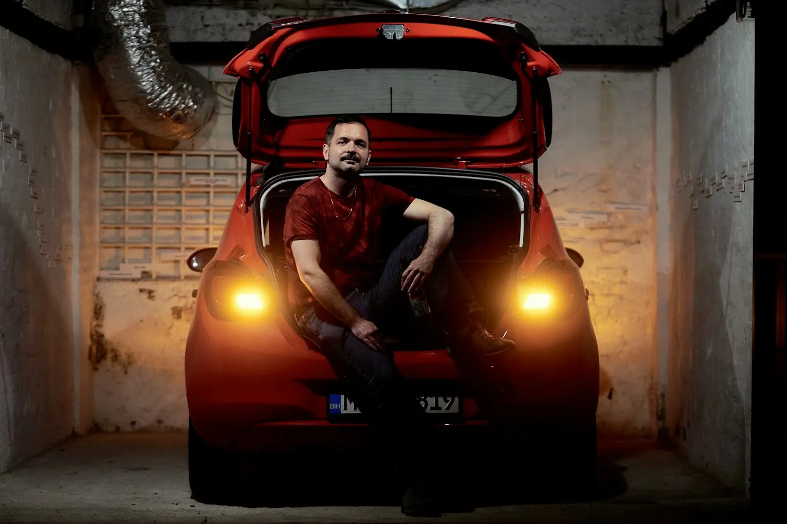 Man wearing red t-shirt and dark panks sitting in the trunk of red car in a garage, bojan stojcic 402 kilometers: objects in the mirror, photo documentation of performance.