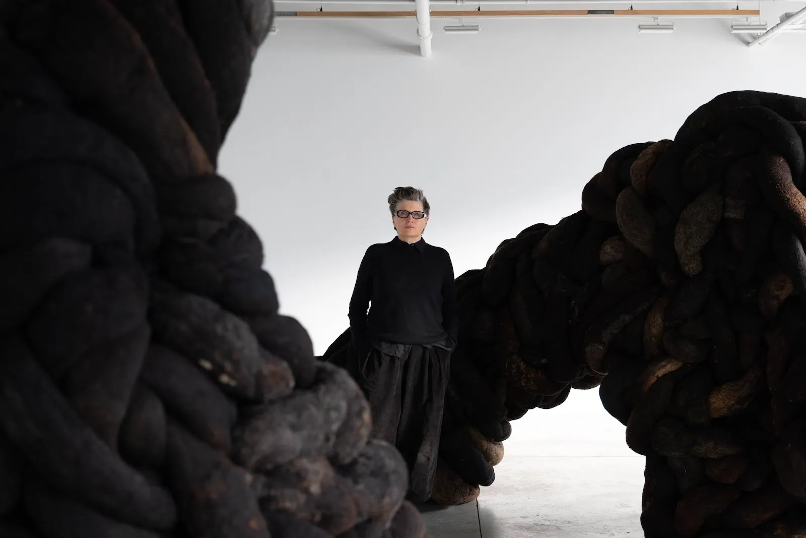 An artist wearing all black stands amid her room-sized sculptural installation, fabric of undoing, in which horsehair and wool form large tendrils coiling around gallery room in carvalho park.
