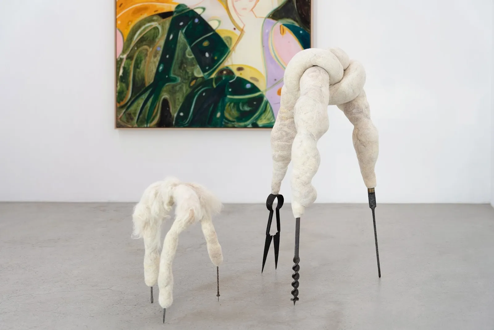 A colorful painting with geometric forms on wall and two sculptures of sheep-like coils mounted on black wooden legs, installation view of the threshold beckons, yulia iosilzon and nicola turner at carvalho park, new york.