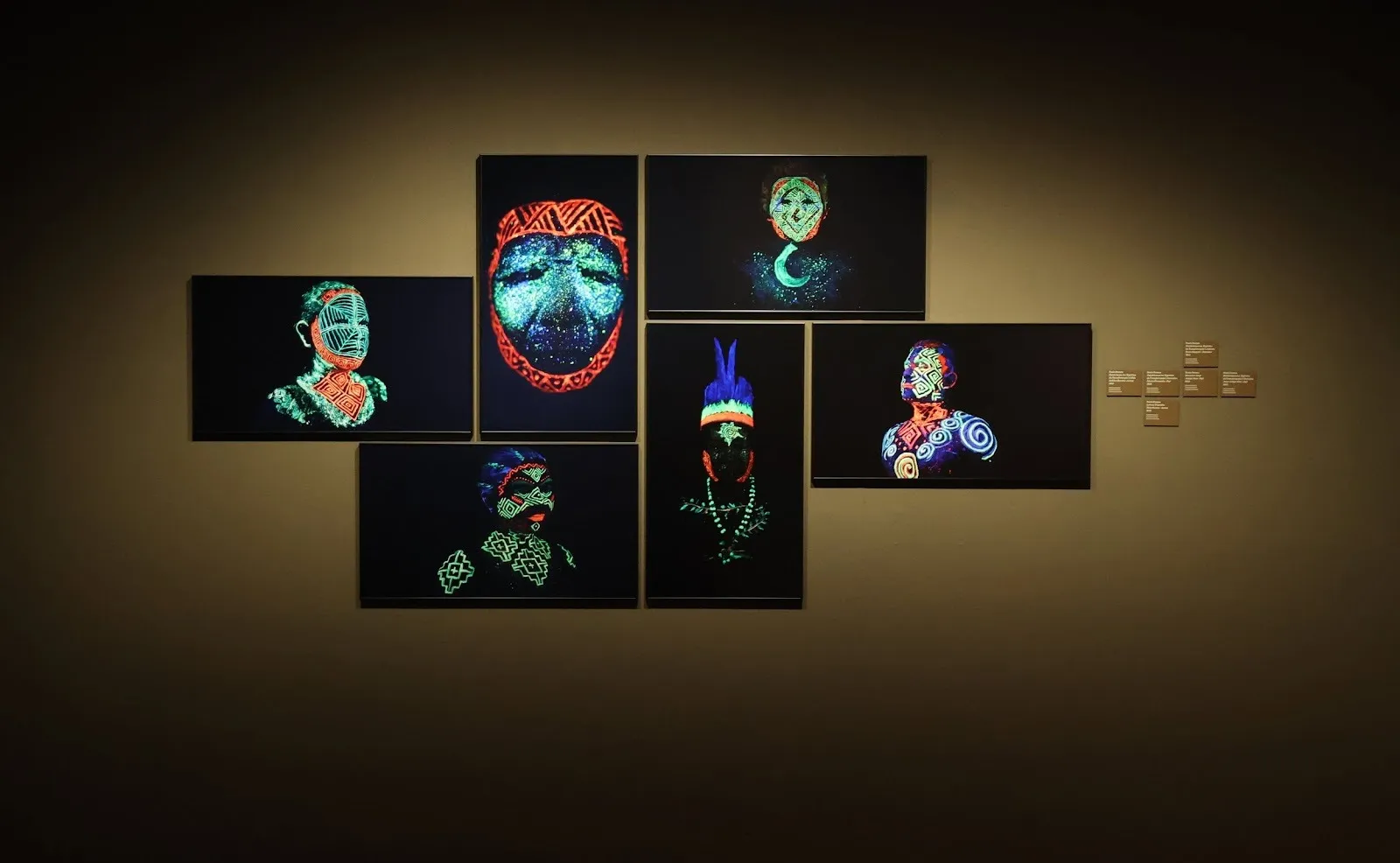Earth-colored wall with patterned renderings of indigenous masks and busts in neon colored lines, installation view of amazons, the ancestral future at the contemporary center of culture in barcelona, CCCB group exhibition.