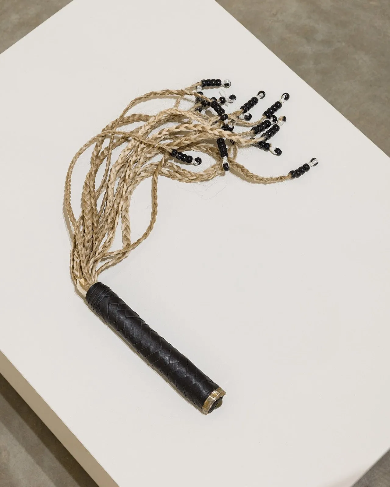 A whip with blond hair braids and black circular beads at the end, sculptural work installed on white pedestal, calli roche all inclusive on view as part of care condition control curated by ae chapman at 601artspace.