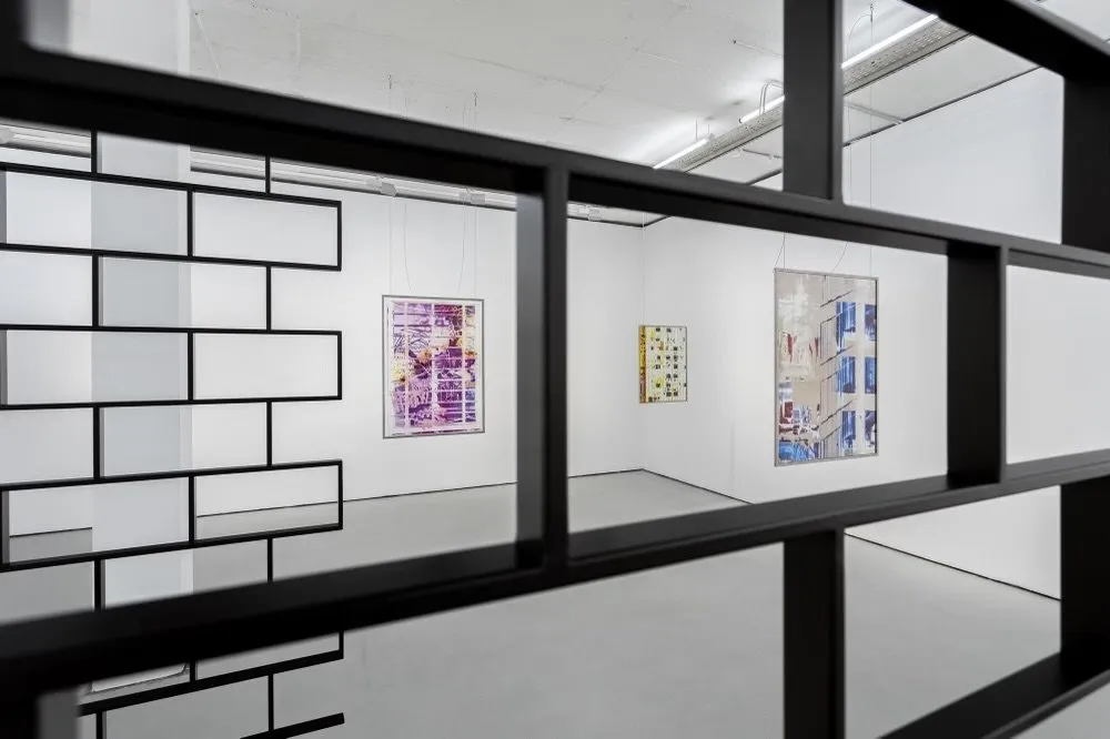 Camera shot looking through a sculptural work of black metal configured in a brick pattern. Through the metal bars, three hanging frames of translucent, colorful images and media layered on top of each other can be seen in the background.