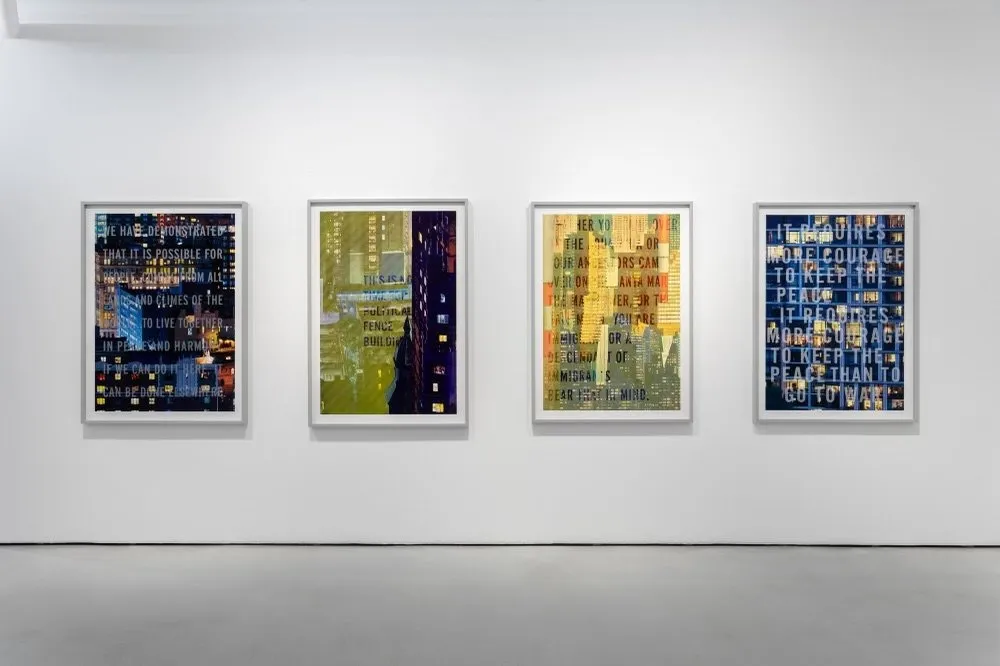 Four frames which appear the same size hang on a single white wall against a concrete floor. Each features colorful images of cityscapes and buildings, layered on top of each other. Also superimposed upon the images are blocks of text.