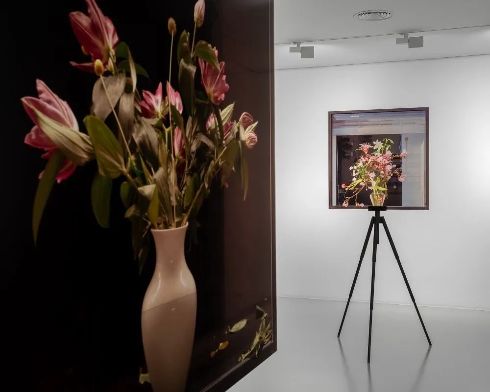 Installation shot of a white cube gallery room. Close to the camera on the left side is an image of flowers in a vase against a dark background. Farther away, there's a frontal view of a similar photograph of flowers, the frame resting upon a tripod.