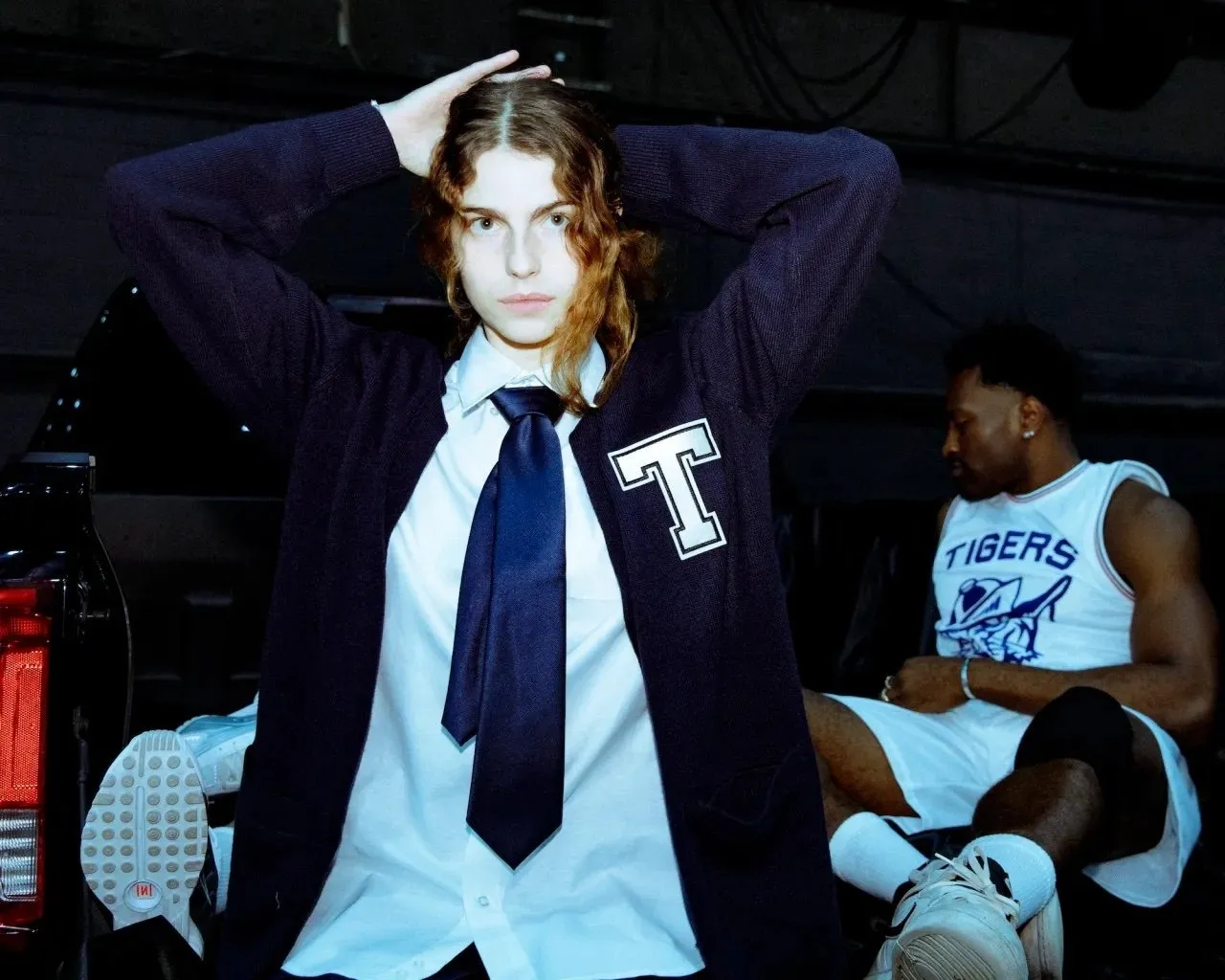 A woman wearing white shirt Navy tie, and cardigan with the letter T staring at the camera, a man in basketball jersey sitting behind her with "tiger" lettering, perla haney jardine in anne imhof's doom house of hope at park avenue armory.