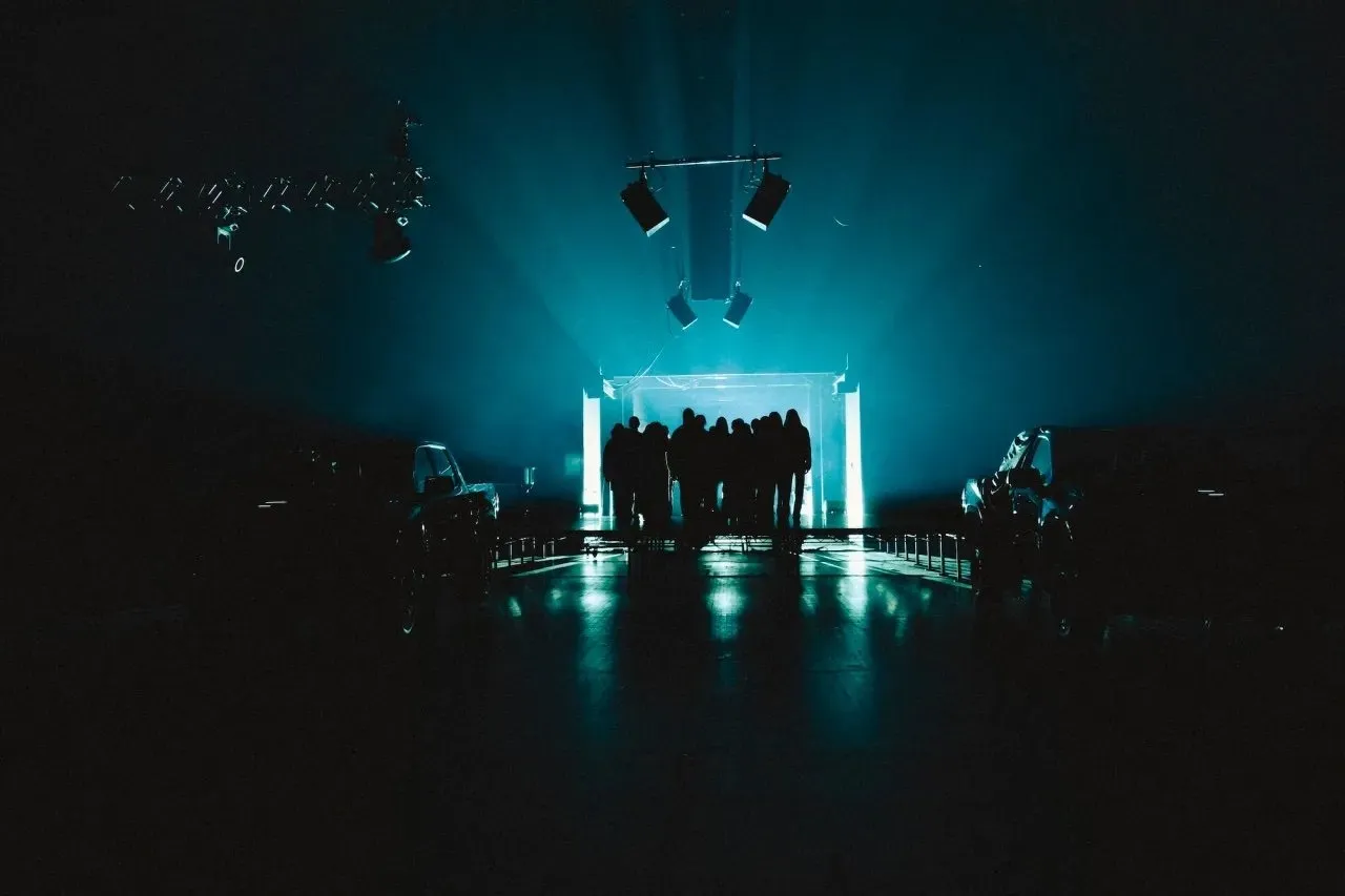 Performers congregate in front of a door with futuristic blue lighting in the middle of a stage at  Anne Imhof’s “DOOM: House of Hope” at Park Avenue Armory.