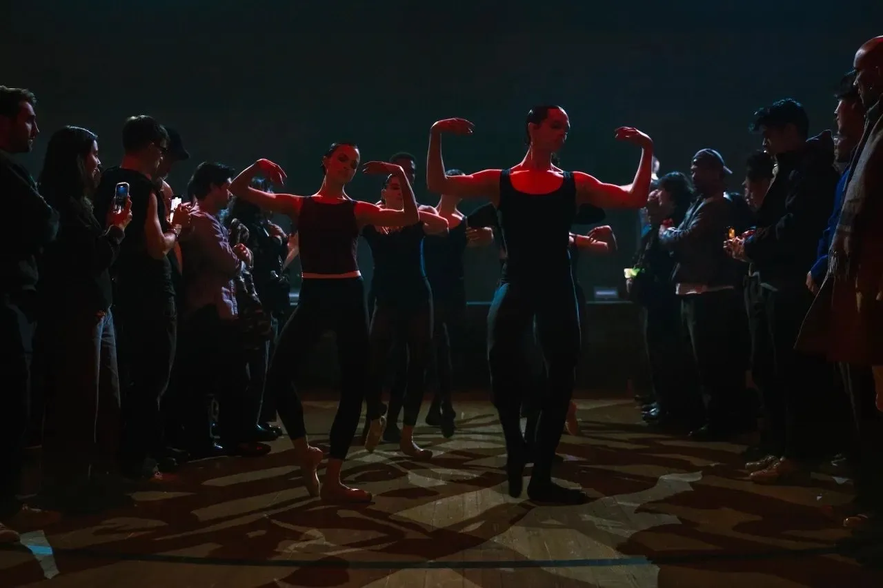 Ballet dancers standing in the crowd performing with their arms vertical to the body under dark lighting, Anne Imhof’s “DOOM: House of Hope” at Park Avenue Armory.