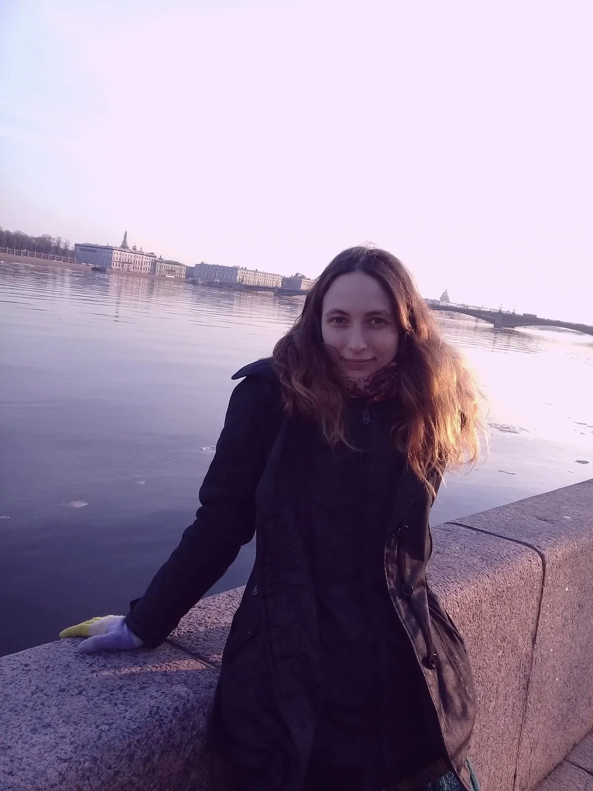 Photo of artist and illustrator Sasha Skochilenko in black jacket leaning against a wall next to river.
