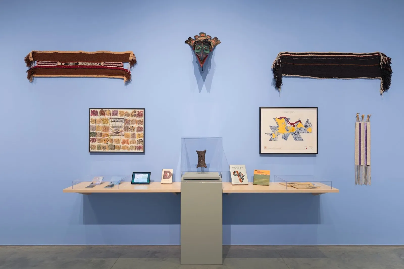 Folk traditional objects, a mask, collages, and design work installed on blue wall at ford foundation gallery, reverberations lineages in design history installation view.