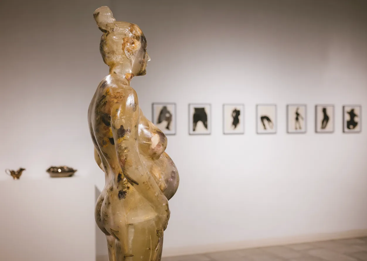 Soap sculpture of pregnant woman with bullets and flowers embedded, in the background there are watercolors of female bodies and vessels, maria kulikovska once lega found an egg blue like a hyacinth, exhibition view at mriya gallery.