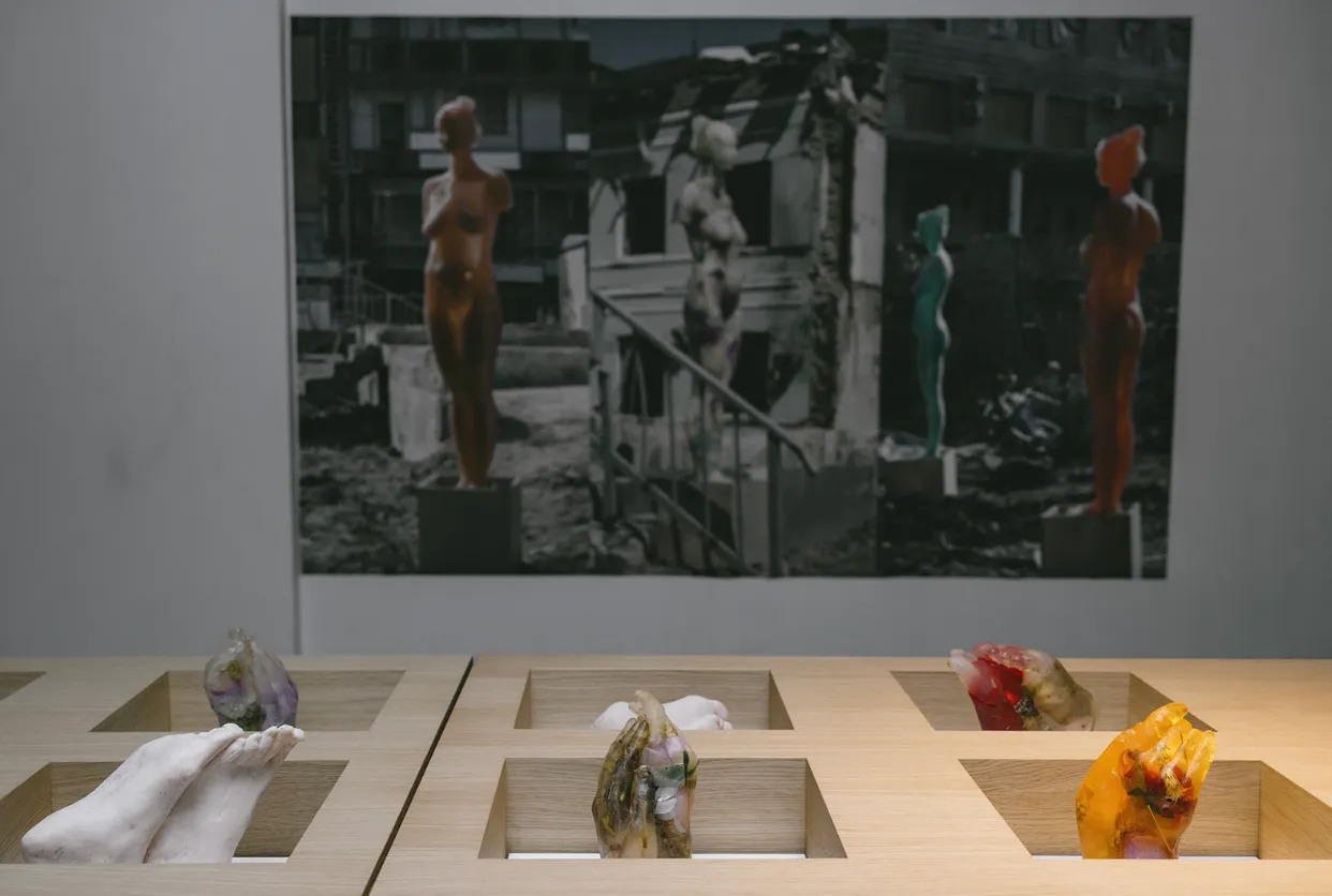 Six hand and feet soap sculptures with embedded flowers displayed on wooden table, with photograph in the background showing female bodies in architectural remains, maria kulikovska once leda found an egg blue like a hyacinth solo exhibition.