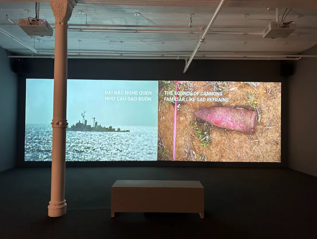 Installation shot of 2 floor-to-ceiling screens. Left screen features a military ship out on sea with text "Đại Bác Nghe Quen Như Câu Dạo Buồn." Right screen features a bomb in the dirt with text "The sound of canons familiar like sad refrains."