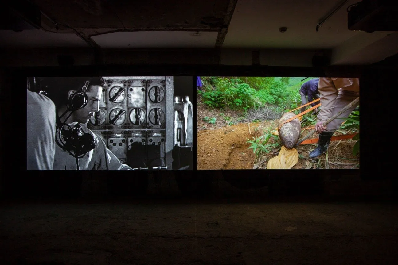 Installation shot of a dark room with two large, floor-to-ceiling screens. The screen on the left features a black and white image of a man inside a radio communications room. The right screen as a color image of a bomb being excavated from dirt.