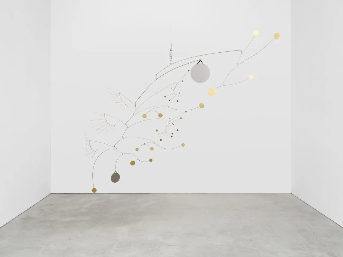 Installation shot of gallery room with white walls and concrete floor. Hanging from the ceiling is a mobile sculpture of thin metal branches hanging from each other, supporting silver and metal discs of various sizes at their ends.
