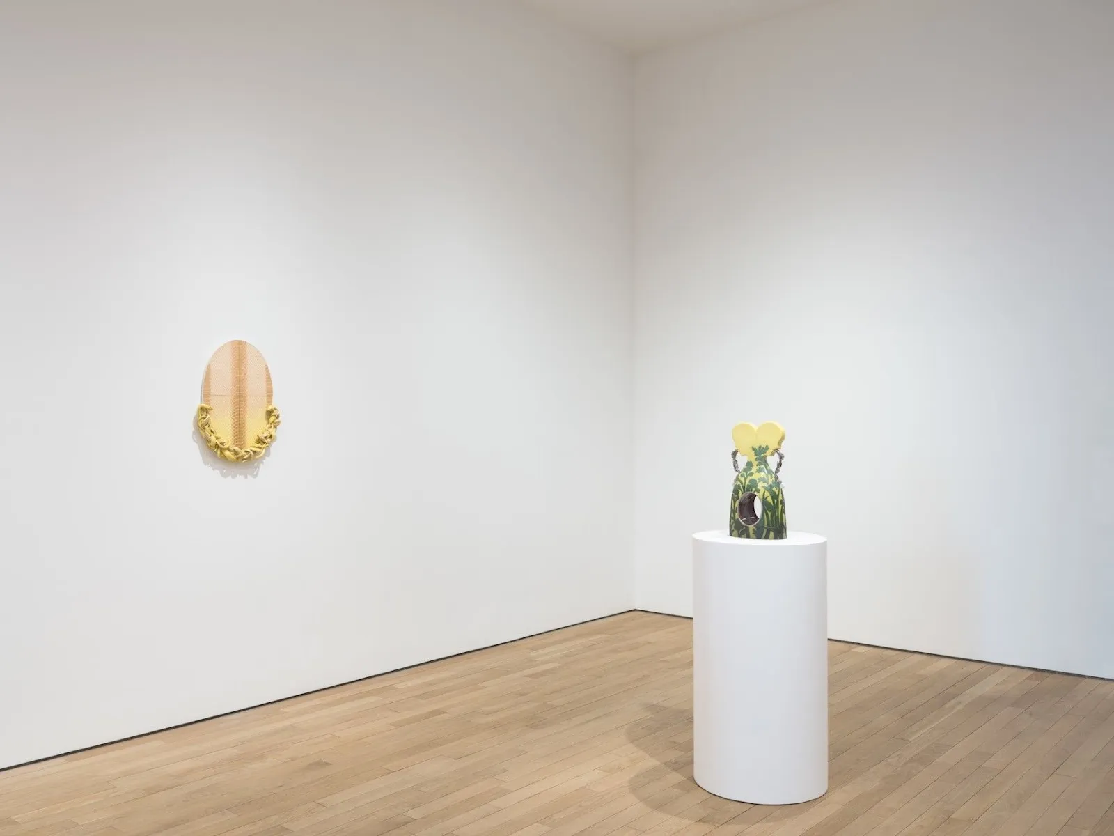 Installation corner shot of gallery with white walls and hardwood floors. The left wall features a flat oval sculpture with ornamentation on bottom. Towards the right is a circular plinth supporting a yellow and green sculpture.