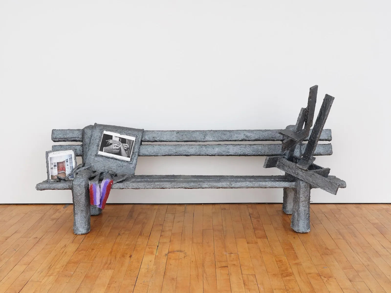 Installation shot of white gallery wall and hardwood floor. Central and frontal is a sculpture of a gray bench with a textures, metallic surface. The bench is slightly abstracted with various other forms resting upon and coming out of it.