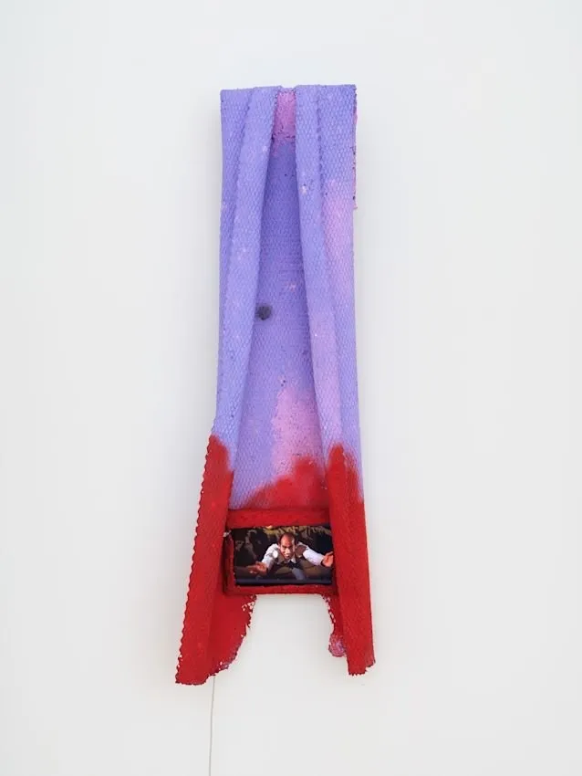 Shot of a hanging piece made from a fabric-like material, dyed lilac and red and folded in on itself. Towards the bottom part of the piece, the fabric frames and reveals a small screen featuring a man seeming to dance.