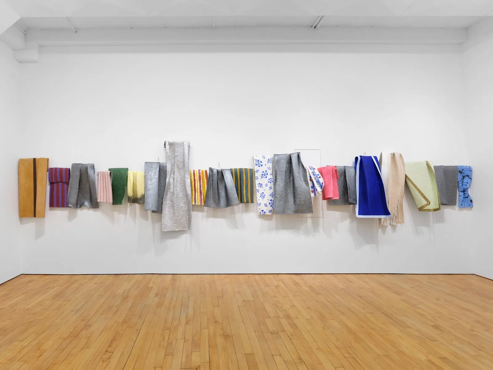 Installation shot of white gallery wall and hardwood floor. Central is a long, horizontal piece mounted to the wall of various textile-like folded rectangular shapes of various color and size.