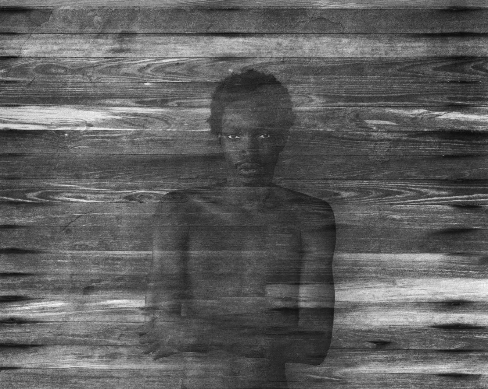 Black and white print of topless young boy looking intently at camera with arms crossed, Tyler Mitchell, Lamine's Apparition (After Frederick Sommer), 2024, photography gelatin silver print.