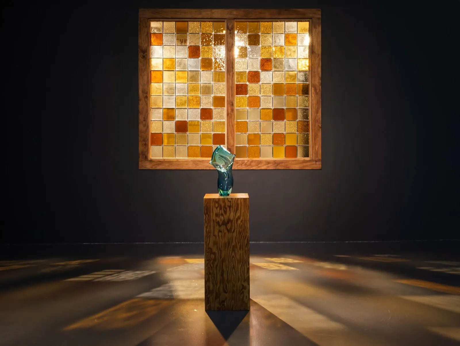 A mosaic stained glass orange and amber window casts light through it, in fron tof a wooden pedestal with blue blown glass sculpture, le'andra leseur exhibition at pittsburgh cultural trust.