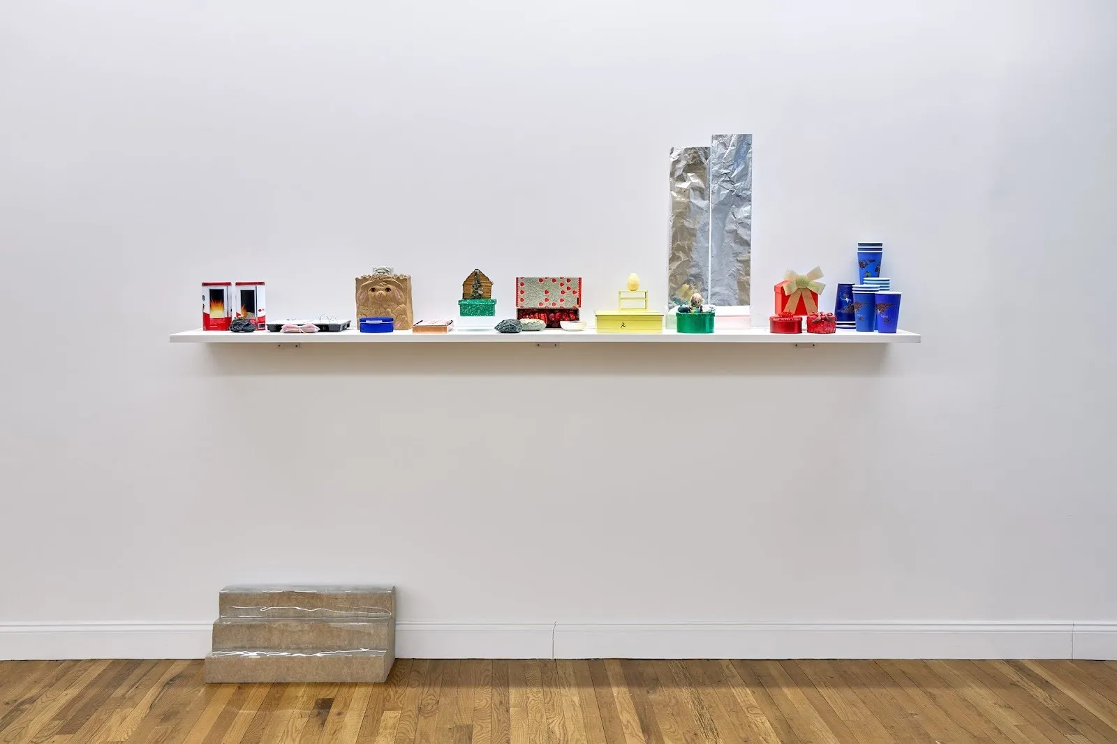 Collected objects placed on a white shelf with stair-shaped stand underneath in front of white wall, adam milner meanwhile assortment, solo exhibition at rainrain gallery.