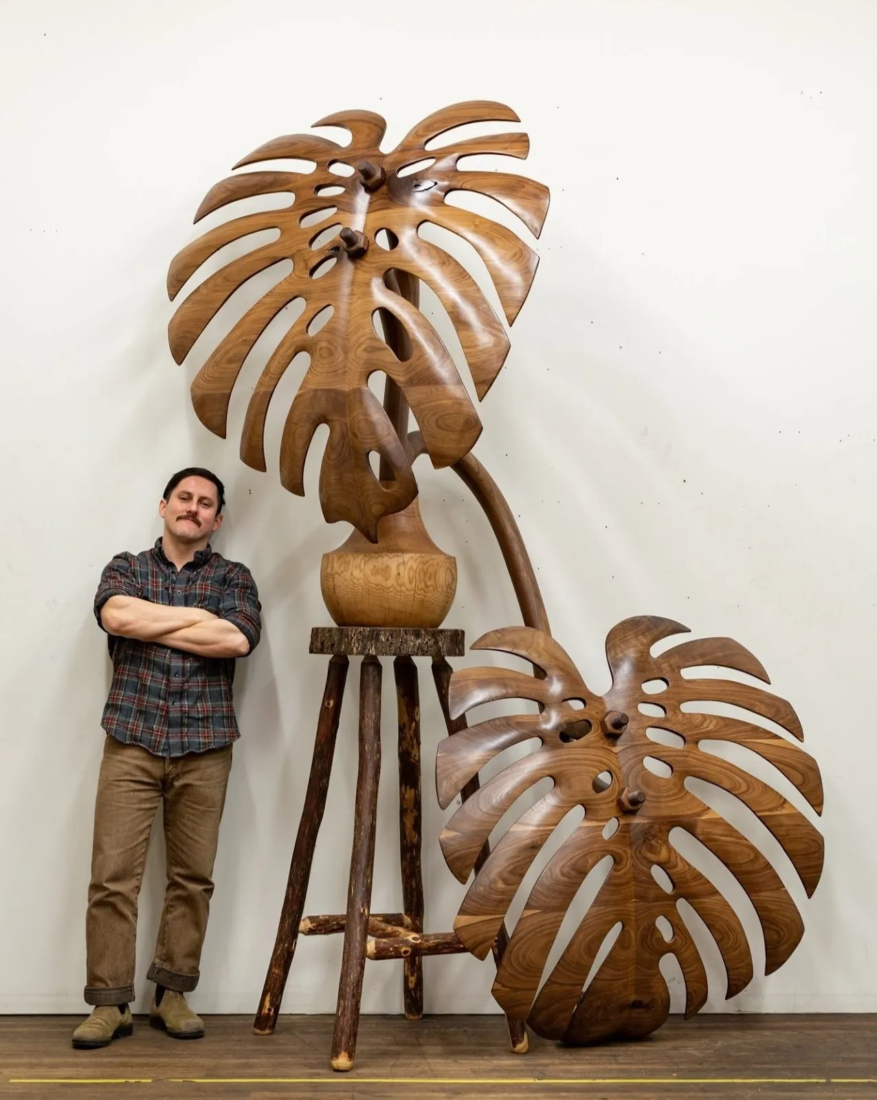 Man in plaid shirt and khaki pants stand next to giant wooden sculpture carved in the shape of a plant on a wooden stool, raul de lara artist portrait.