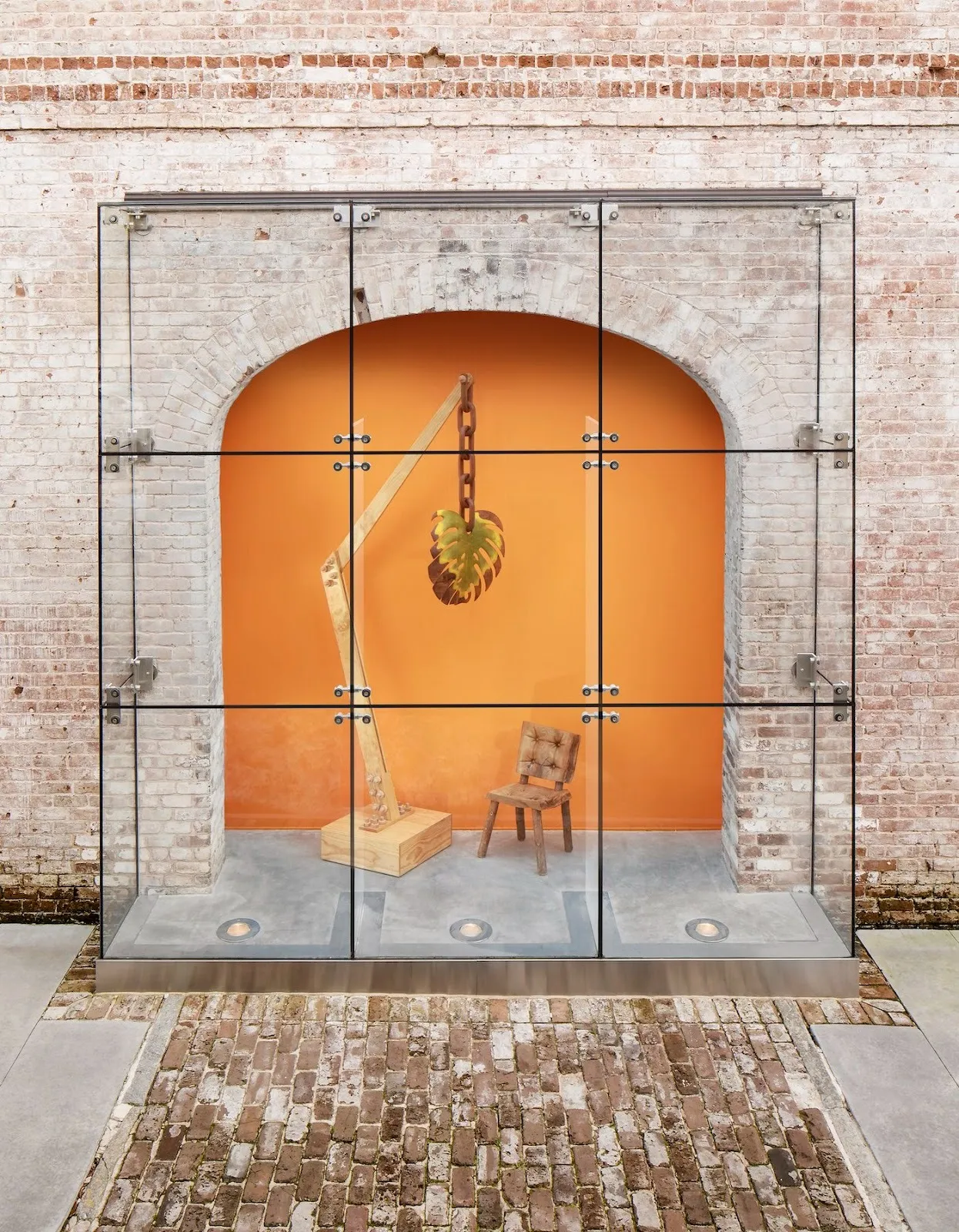 Wooden leaf crane scculpture hanging from a chain next to a chair in front of orange background under an arch, glass case outside, raul de lara, raices/roots at scad museum of art.