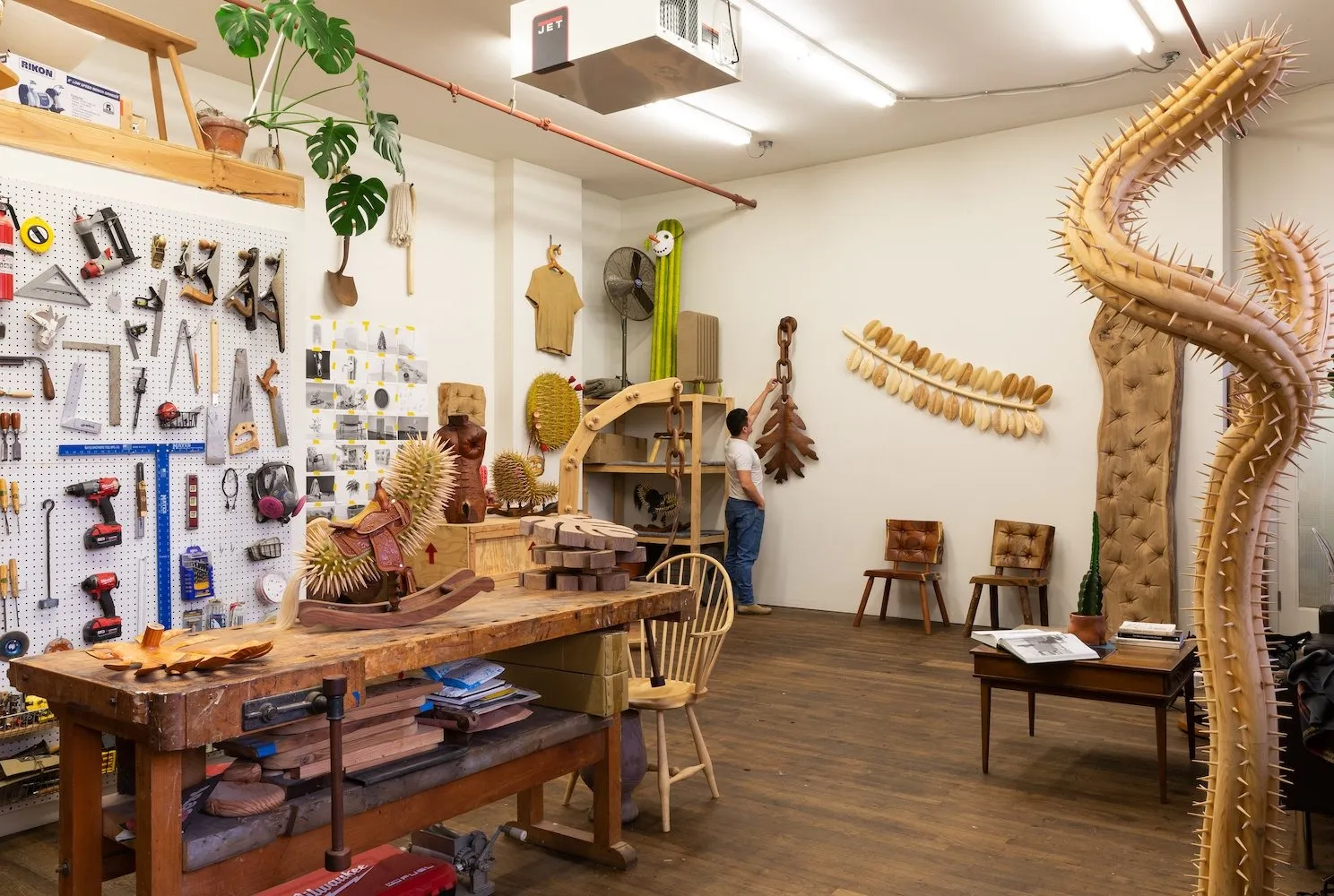 Artist raul de lara in his ridgewood queens studio with woodworking tools hanging on the wall, a few plant sculptures in the middle, and he is fixing a sculpture on the wall.