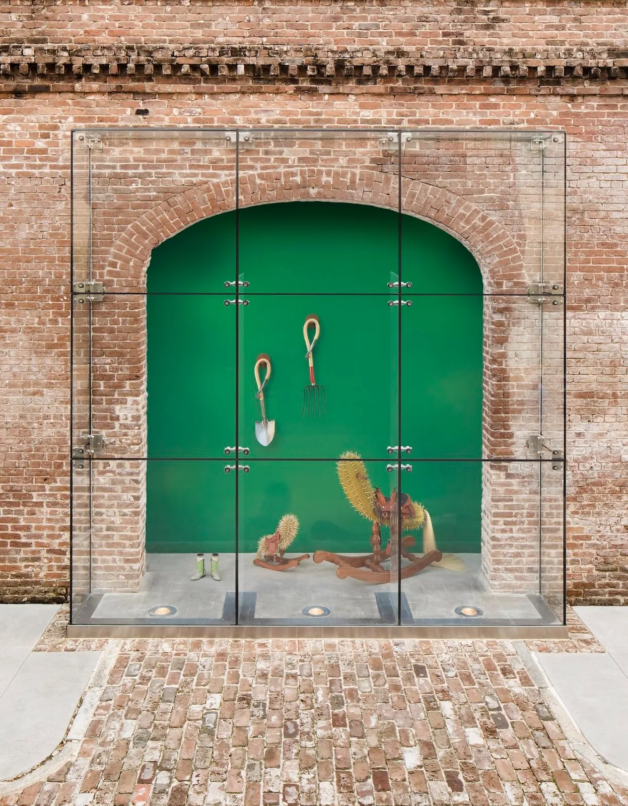 Two cactus wooden sculptures next to two farming tools hanging on green background, a pair of boots displayed on the floor, installation in glass case, raul de lara raices/roots at scad museum of art.