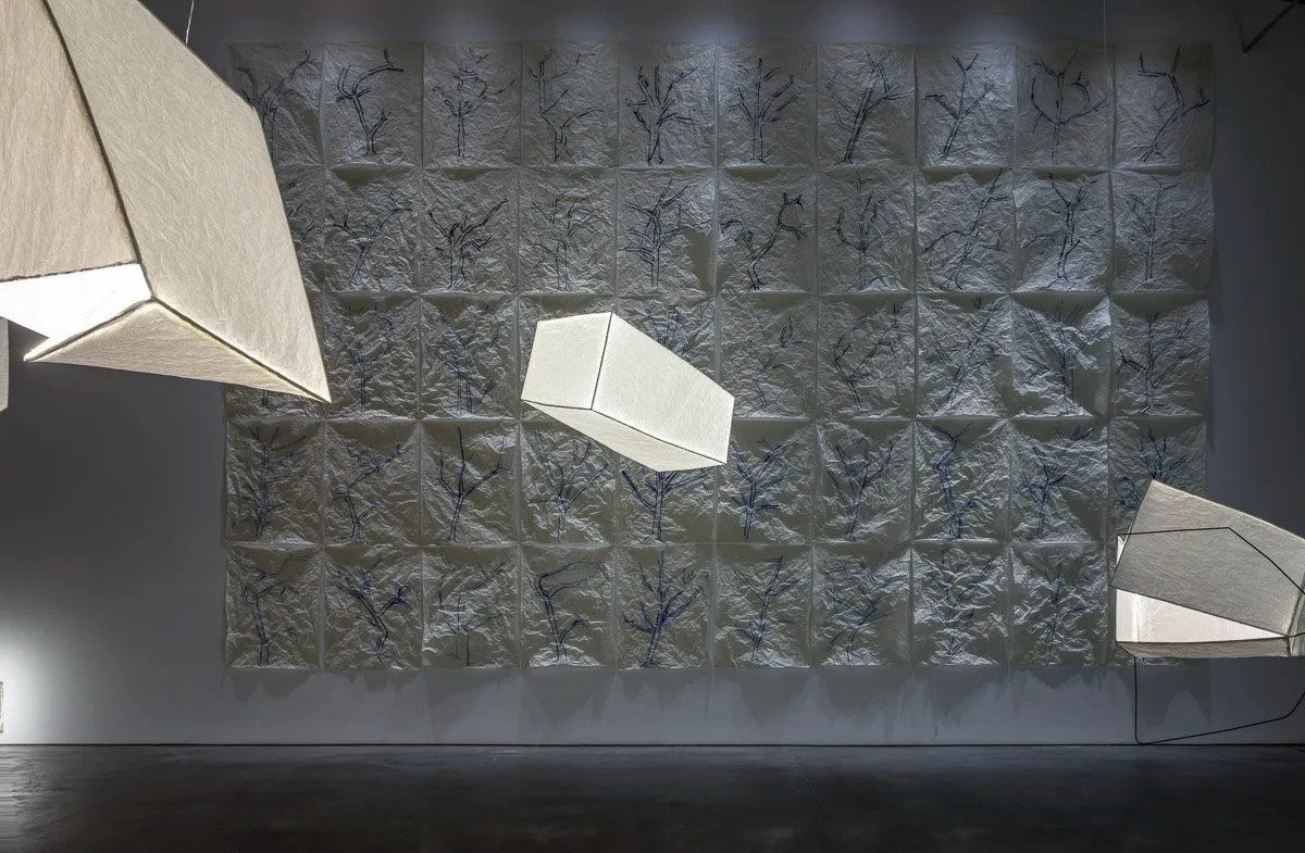 Installation view of dimly lit gallery room with floating hung lantern-like fixtures throughout. In the background, a work in crumpled pieces of paper arranged in a grid feature blue inkings of tree-like shapes.