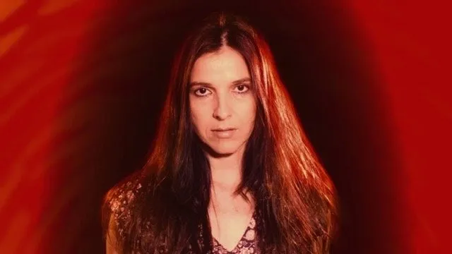 Long-haired woman looking at the camera in front of red background, chronicles of the absurd by miguel coyula and lynn cruz.