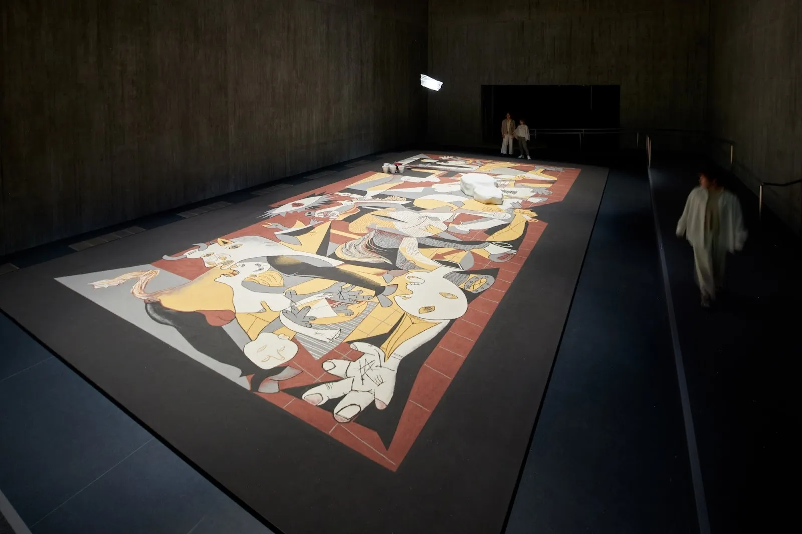 Large artwork by lee mingwei titled guernica in sand lying horizontally, recreation of picasso's work against dark background inside gallery of M+, Hong Kong.