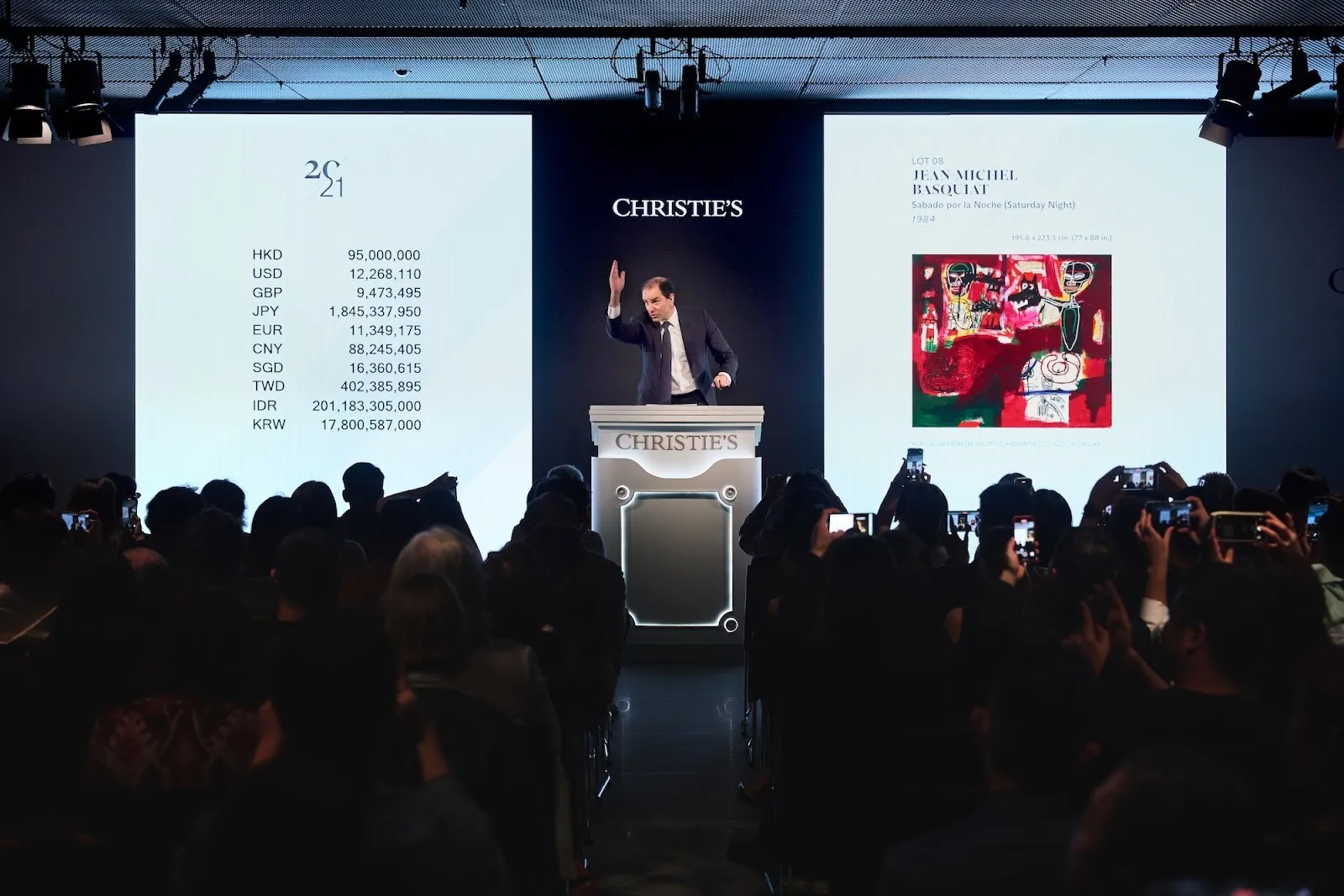 In front of two projection screens with artwork image and prices, an auctioneer stands at podium with hand waving in the air mid-auction, christies hong kong art week.