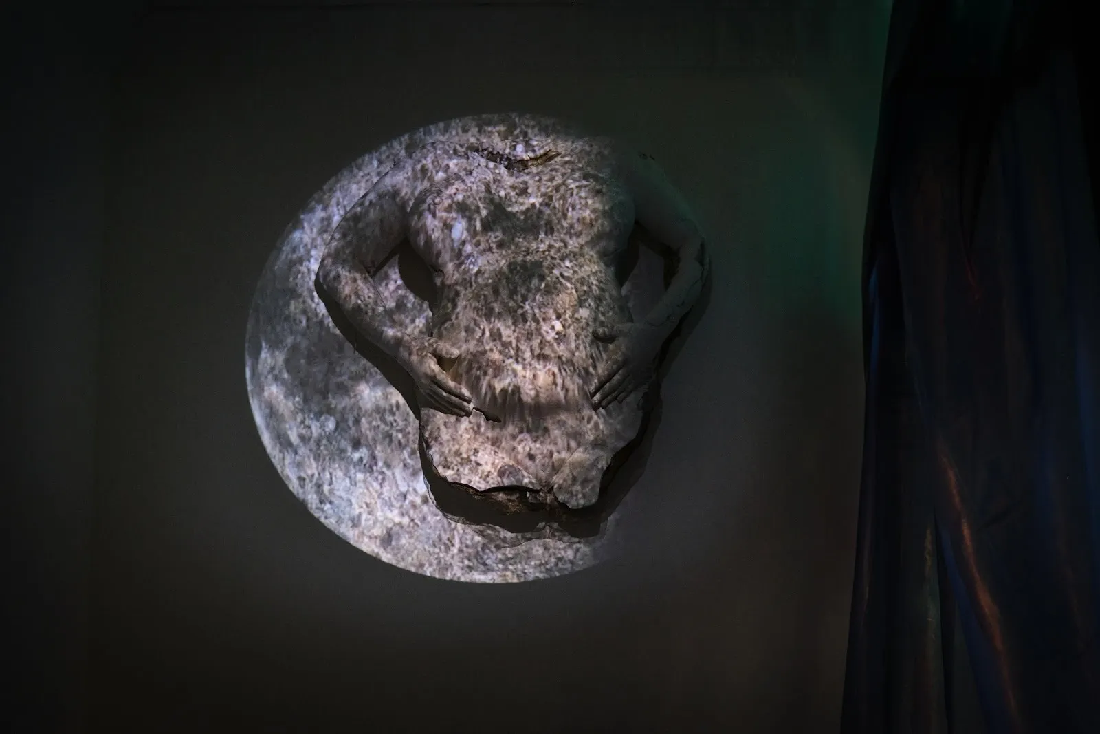 Installation shot of a sculpture of a pregnant torso and arms mounted to a wall. Projected onto the sculpture is a moon-like image.