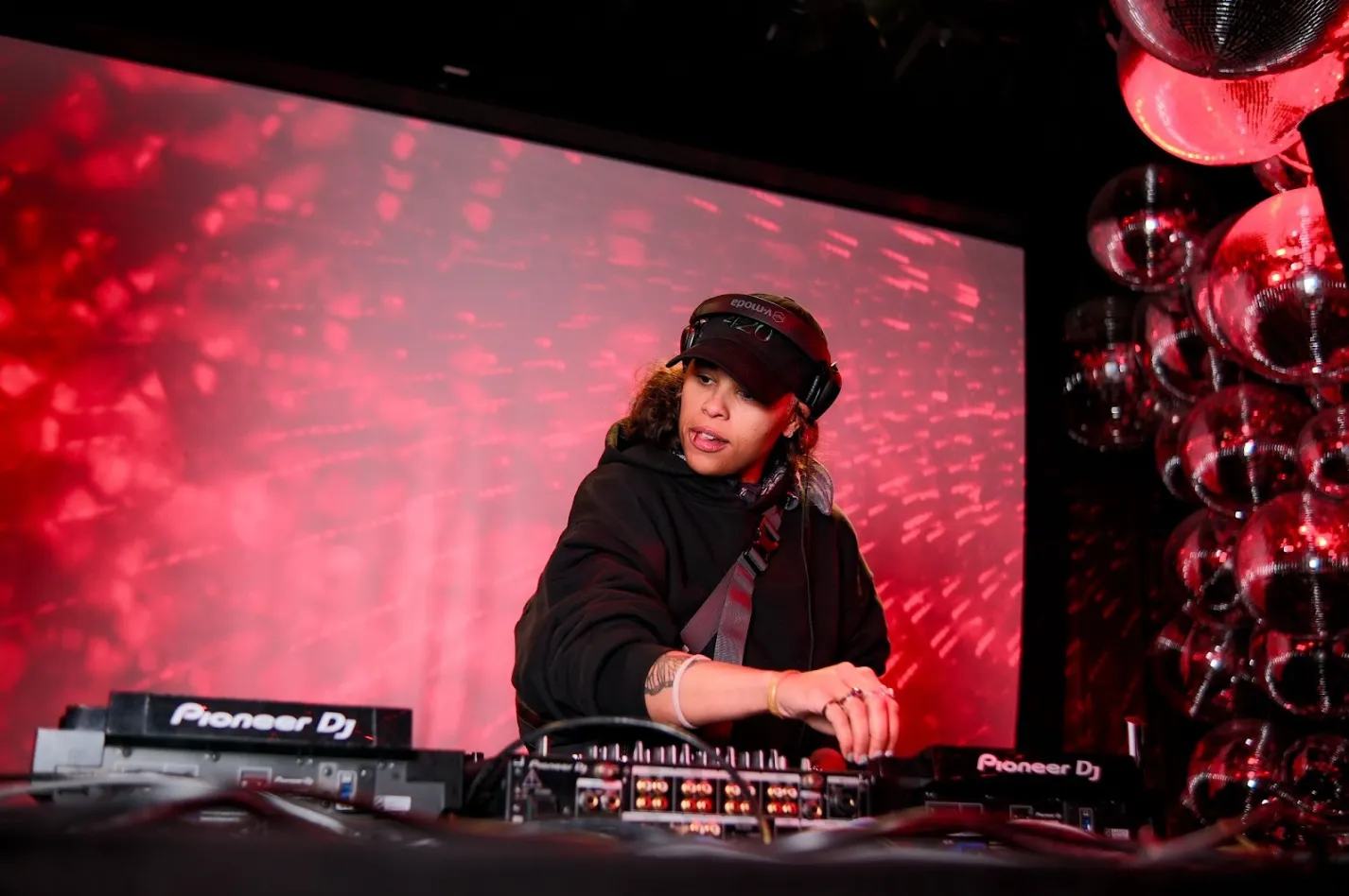 Shot of a DJ performing a set. In the background, a large screen projects blurry red lights and a wall of disco balls towers to the right of the DJ.