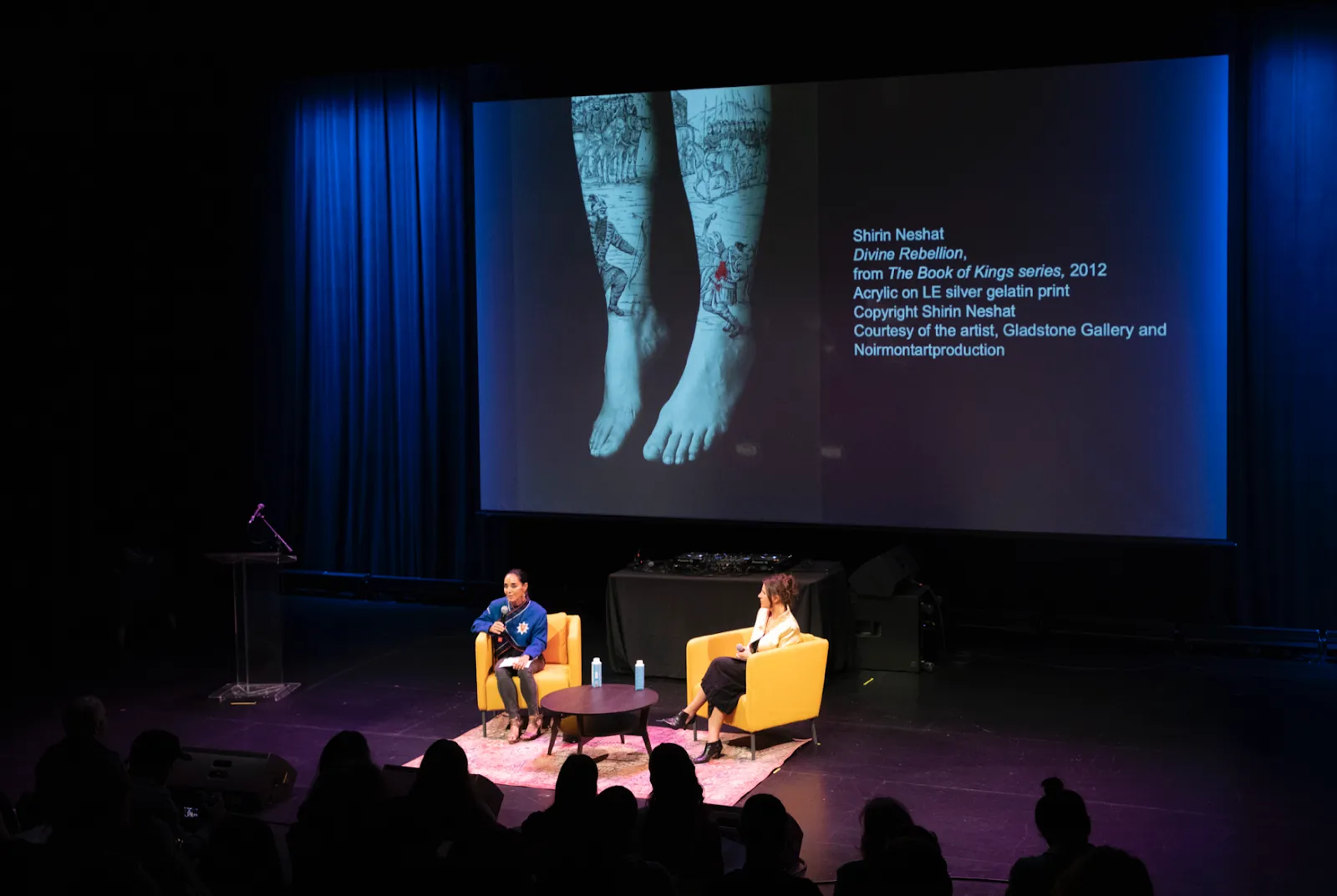 Shot of a dimly-lit auditorium with two figures seated on-stage. The left figure is speaking, while an image is projected behind.