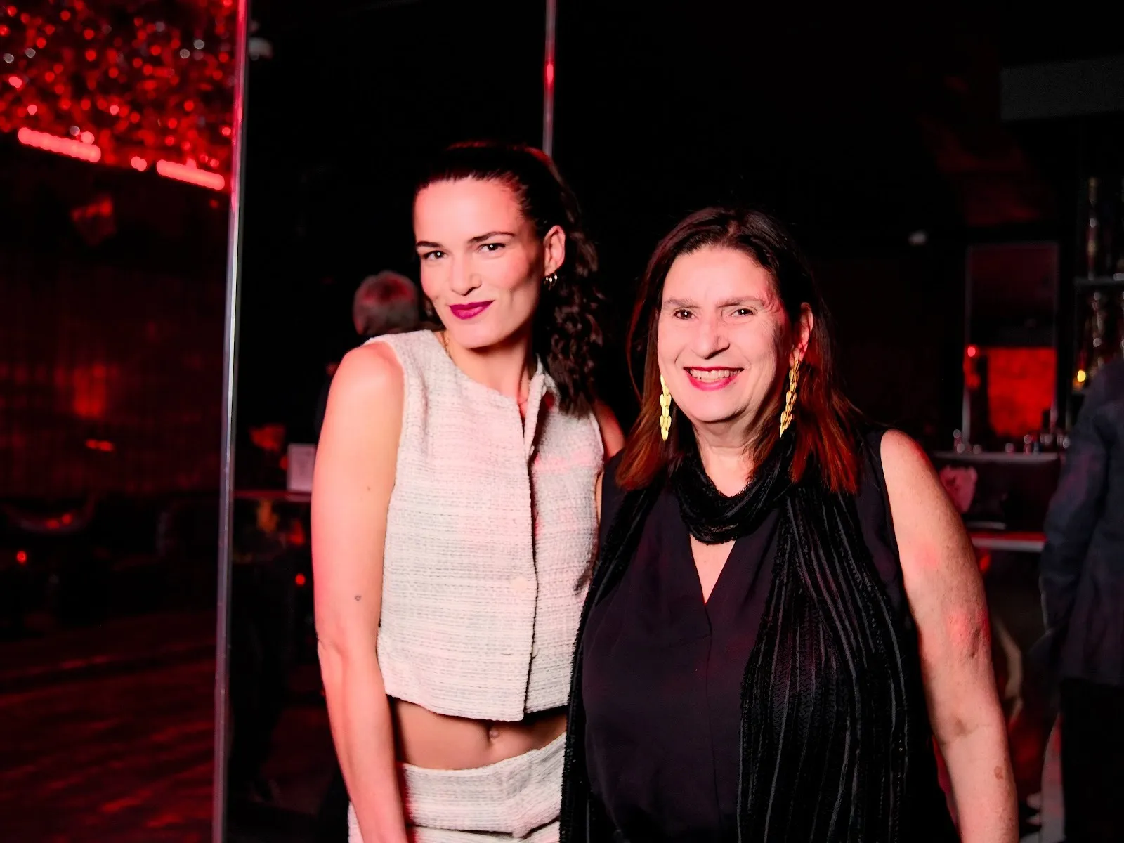 Photo of two women smiling. The woman on the left wears a coordinated, tweed-like white vest and pants set. The woman on the left wears a dark sleeveless tunic and black scarf.