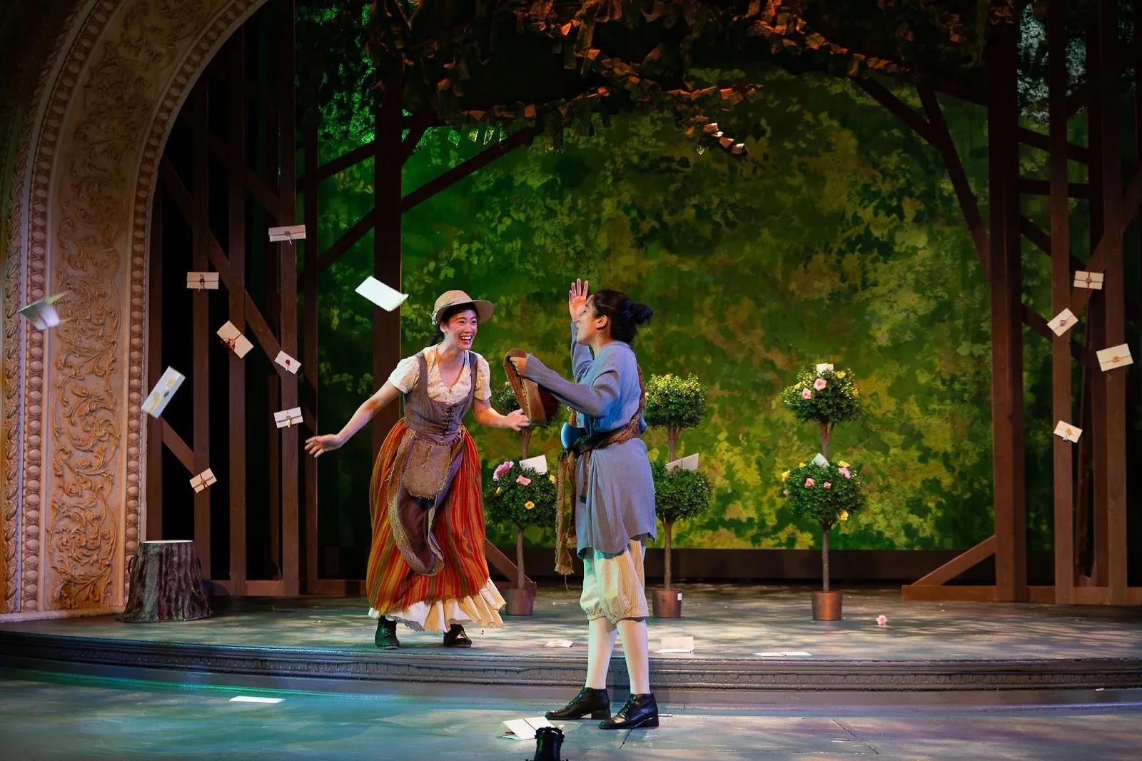 Production photo of Rebecca Ho as Celia in As You Like It, two actors in period costume in front of trees with bills flying around in the air.