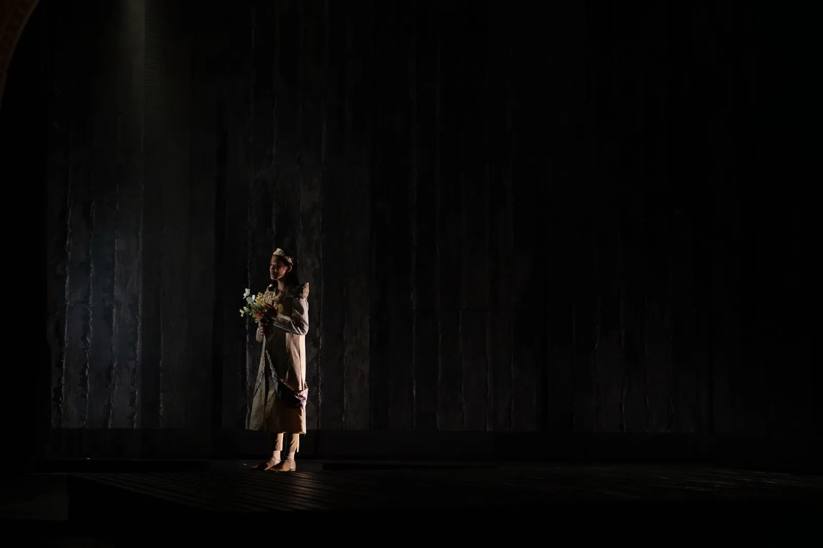 Production photo of Rebecca Ho as Queen in Richard II, dark background with her figure wearing dress and holding a flower bouquet gently contoured on stage.