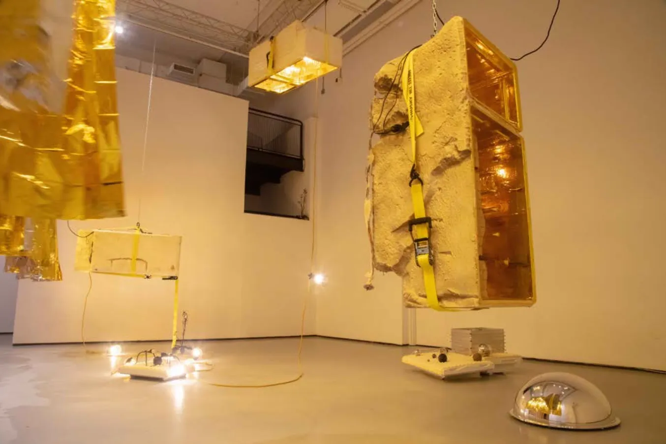 Installation photo from below-up of gallery space with white walls and concrete floor. Large, rough concrete shape and golden metallic material hang from the ceiling. The floor is scattered with various lighting and sculpture.