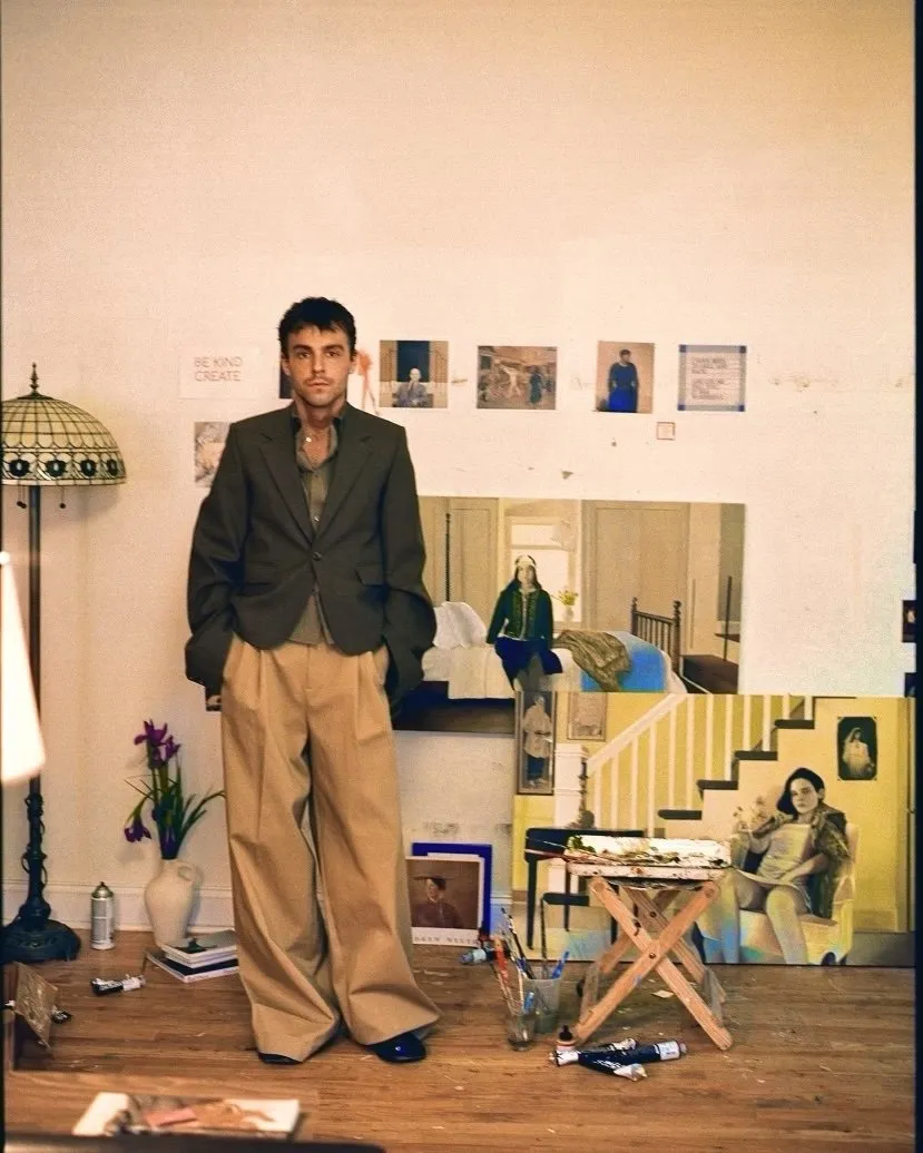 Vintage archival photograph of artist Leonard baby in front of paintings of his sisters, Angels, he's wearing a suit with wide legged pants, hands in pocket staring at camera.