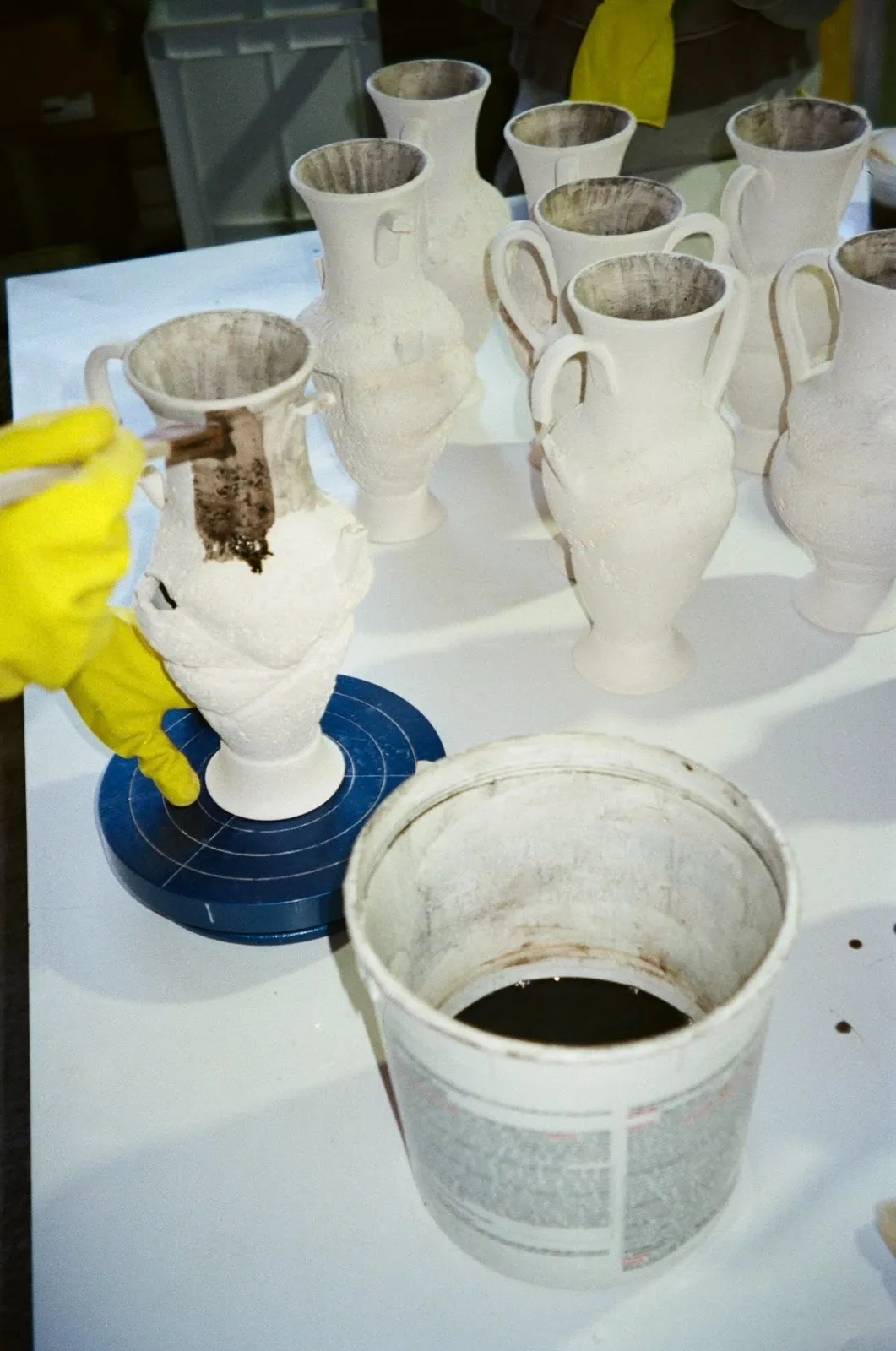 Photo of a gloved hand in motion applying a dark brown glaze to an unfinished amphora. Other unfinished amphorae are clustered in the background on the table.
